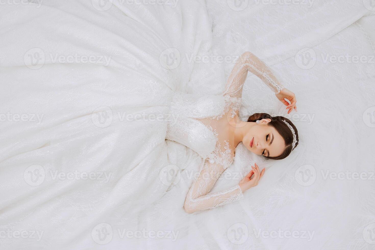 A brunette bride with a tiara in a lacy white dress with long sleeves poses against the background of her dress. On a white background. Classic hairstyle. Evening makeup. Spring wedding photo