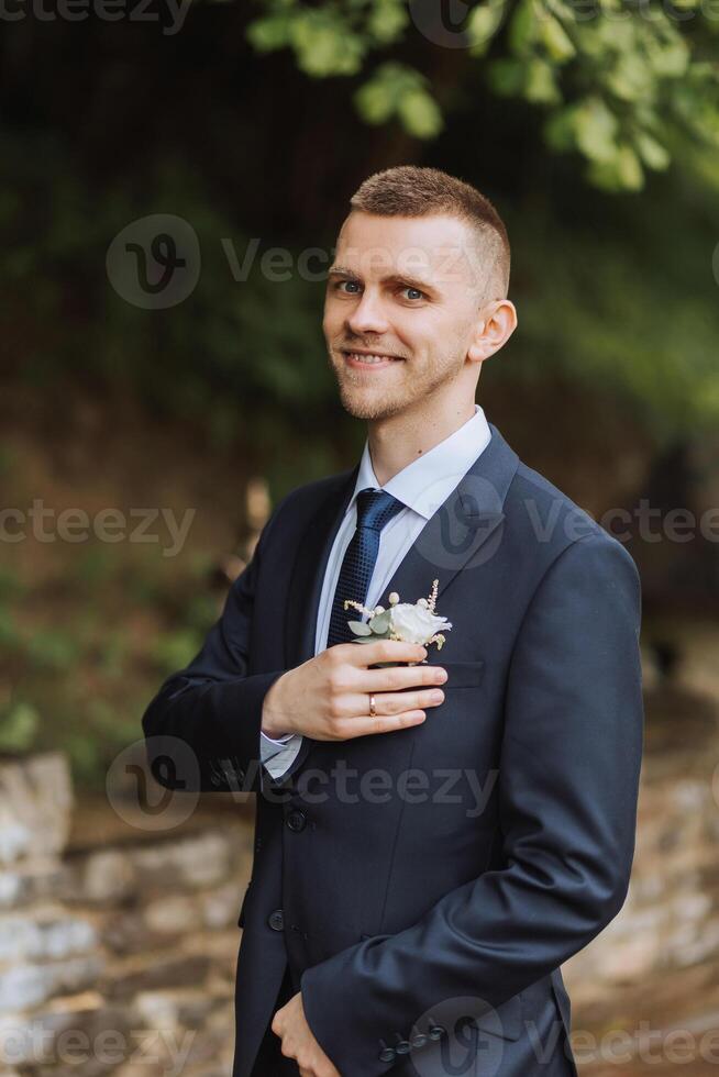 The groom in a black suit adjusts the boutonniere, poses against the background of a green tree. Wedding portrait. photo