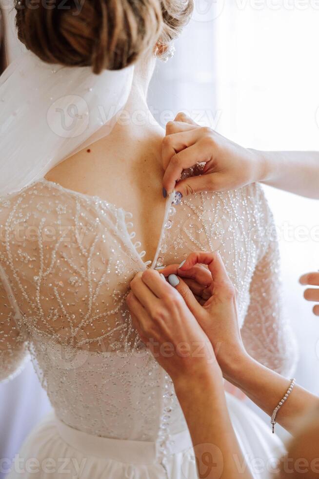 Morning of the bride. The bride's maid of honor helps the bride lace up her dress, fasten buttons on the dress or sleeves. Girlfriends help the bride fasten her dress photo