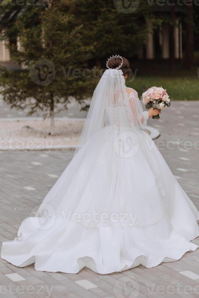 The bride in a magnificent dress and veil walks down the stone path with her shoulders turned. Magnificent dress with long sleeves, open bust. Summer wedding photo