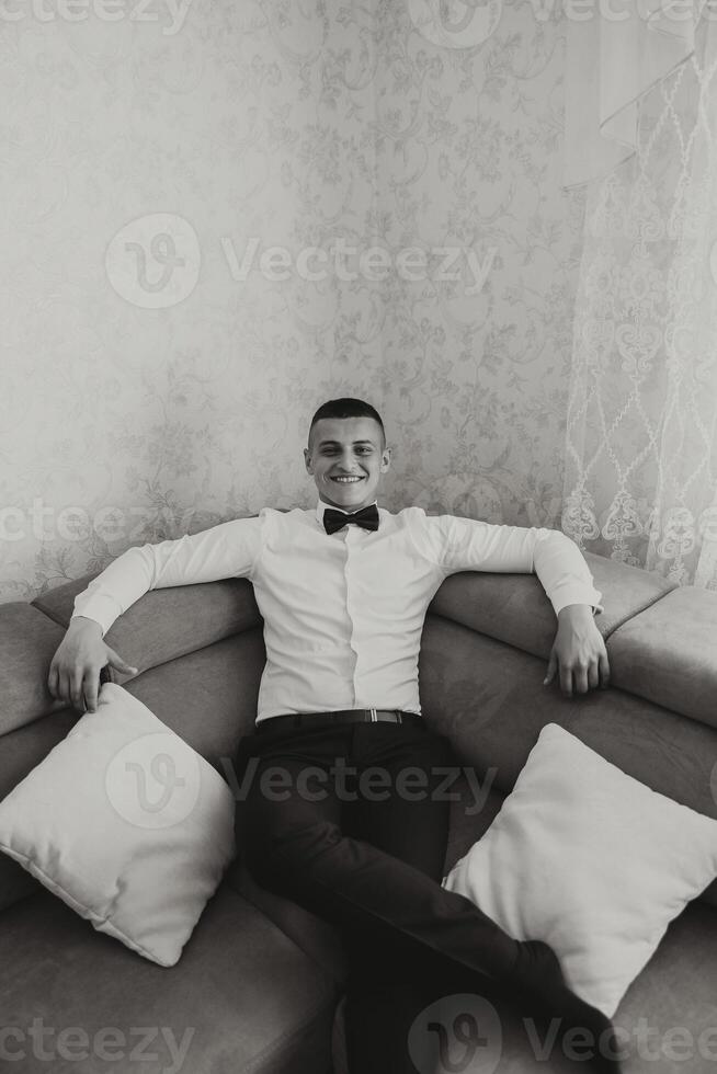 Stylish portrait of the groom preparing for the wedding ceremony. Groom's morning. The groom is sitting on the sofa. The confident look of a mature man. Black and white photo. photo