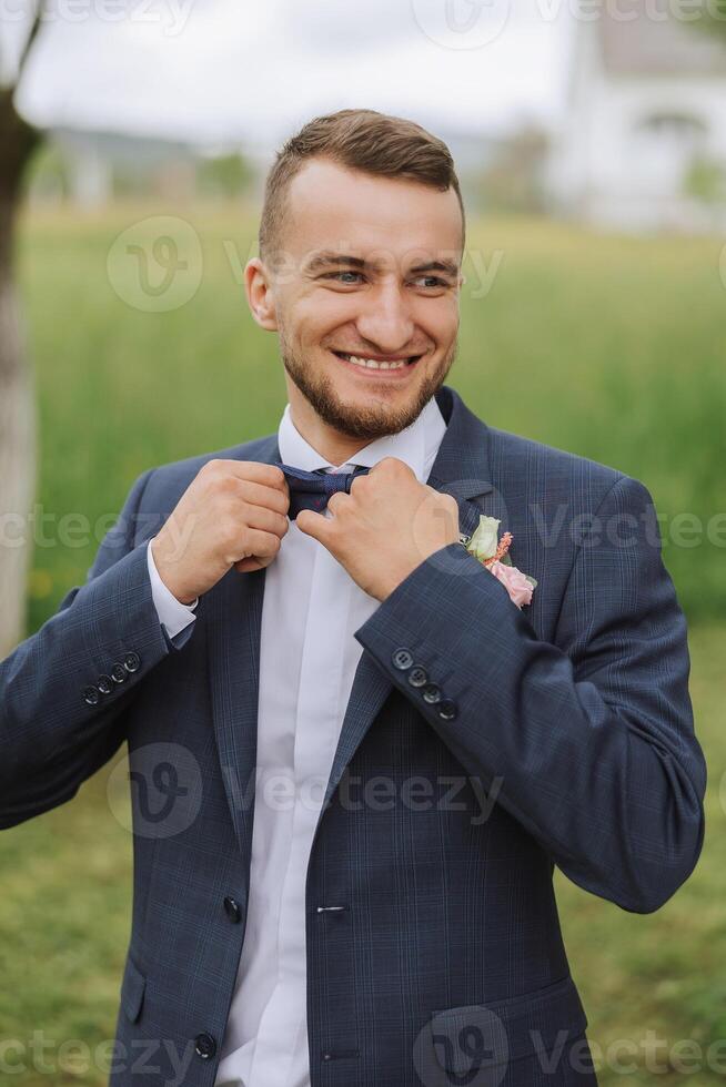 The groom in a black suit adjusts his bow tie, poses against the background of a green tree. Wedding portrait. photo