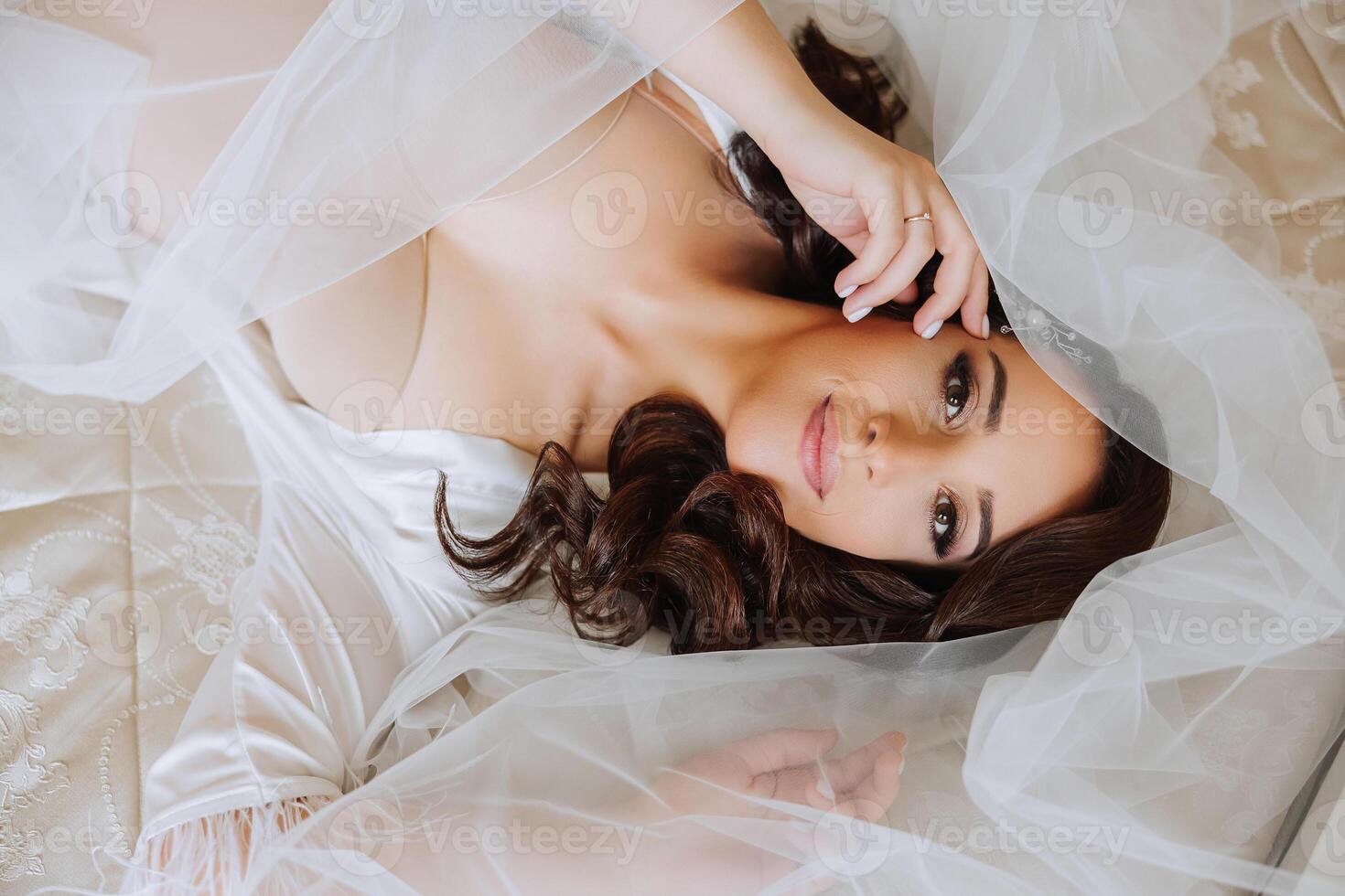 A beautiful brunette bride with a tiara in her hair is preparing for the wedding in a beautiful robe in boudoir style. Close-up wedding portrait of the bride lying on the bed, photo from above.