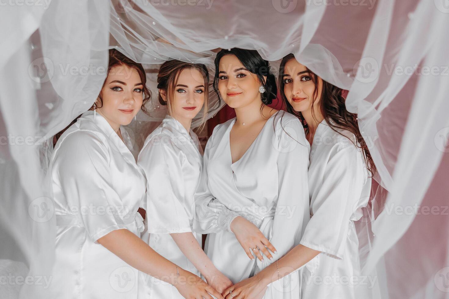 The bridesmaids look at the smiling bride. The bride and her cheerful friends are celebrating a bachelorette party on the bed in identical dresses. Bride and friends in the room. photo