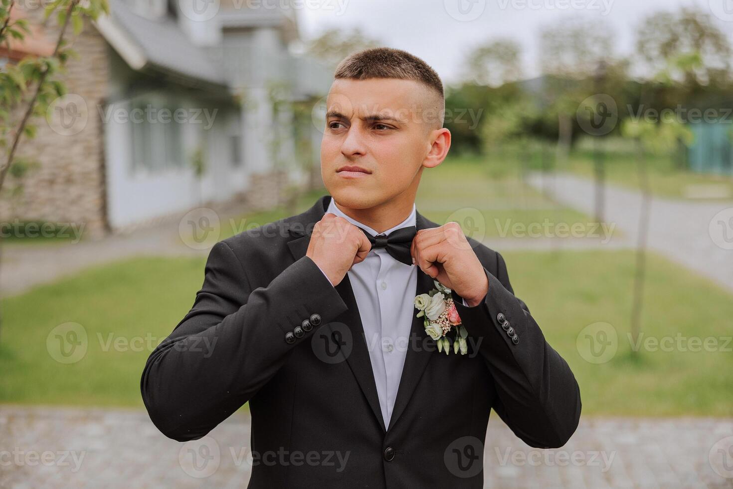 The groom in a black suit adjusts his bow tie, poses against the background of a green tree. Wedding portrait. photo