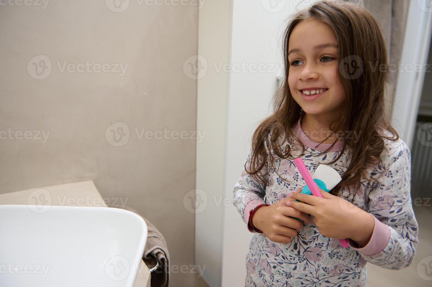 Little child girl in pajamas, holding kids toothpaste and her pink toothbrush, standing in the bathroom at home photo