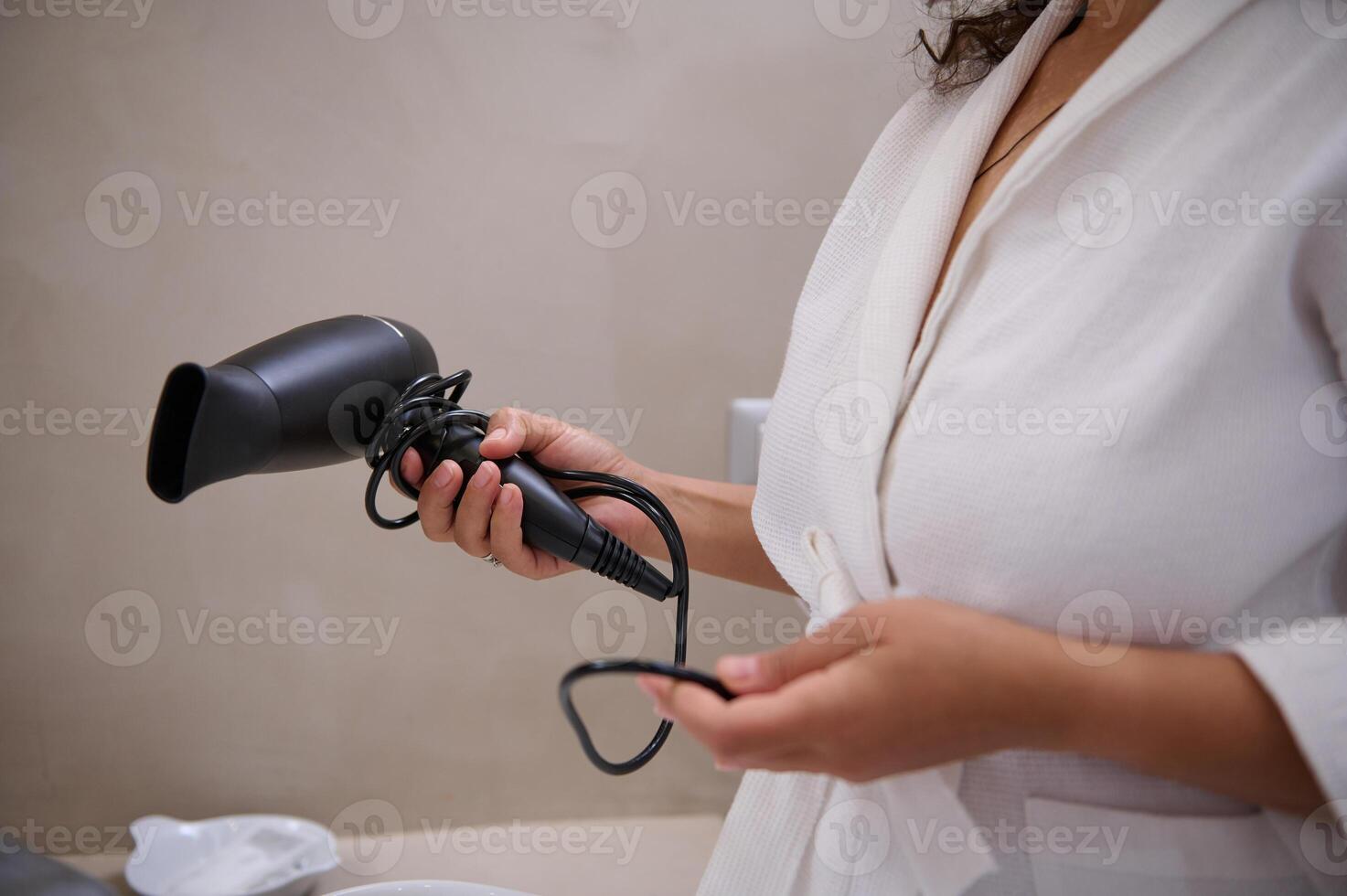 Close-up of a woman in white bathrobe, holding blowing hair dryer, standing in the home bathroom. Copy advertising space photo