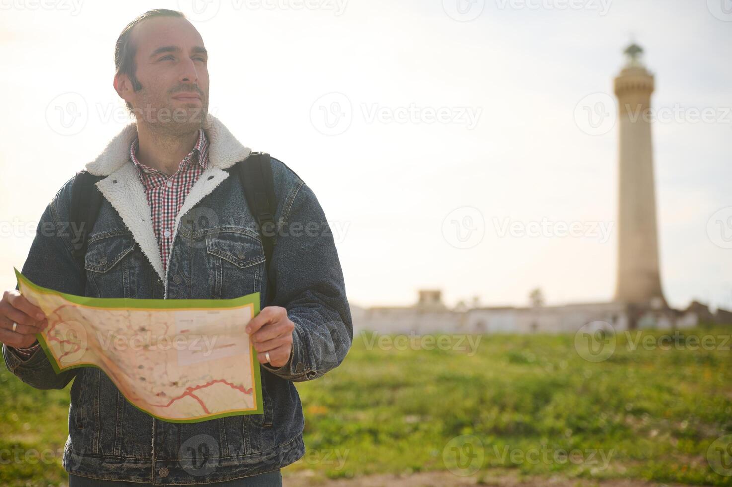 Handsome young male traveler holding a map and looking away, standing on the nature against a lighthouse background photo