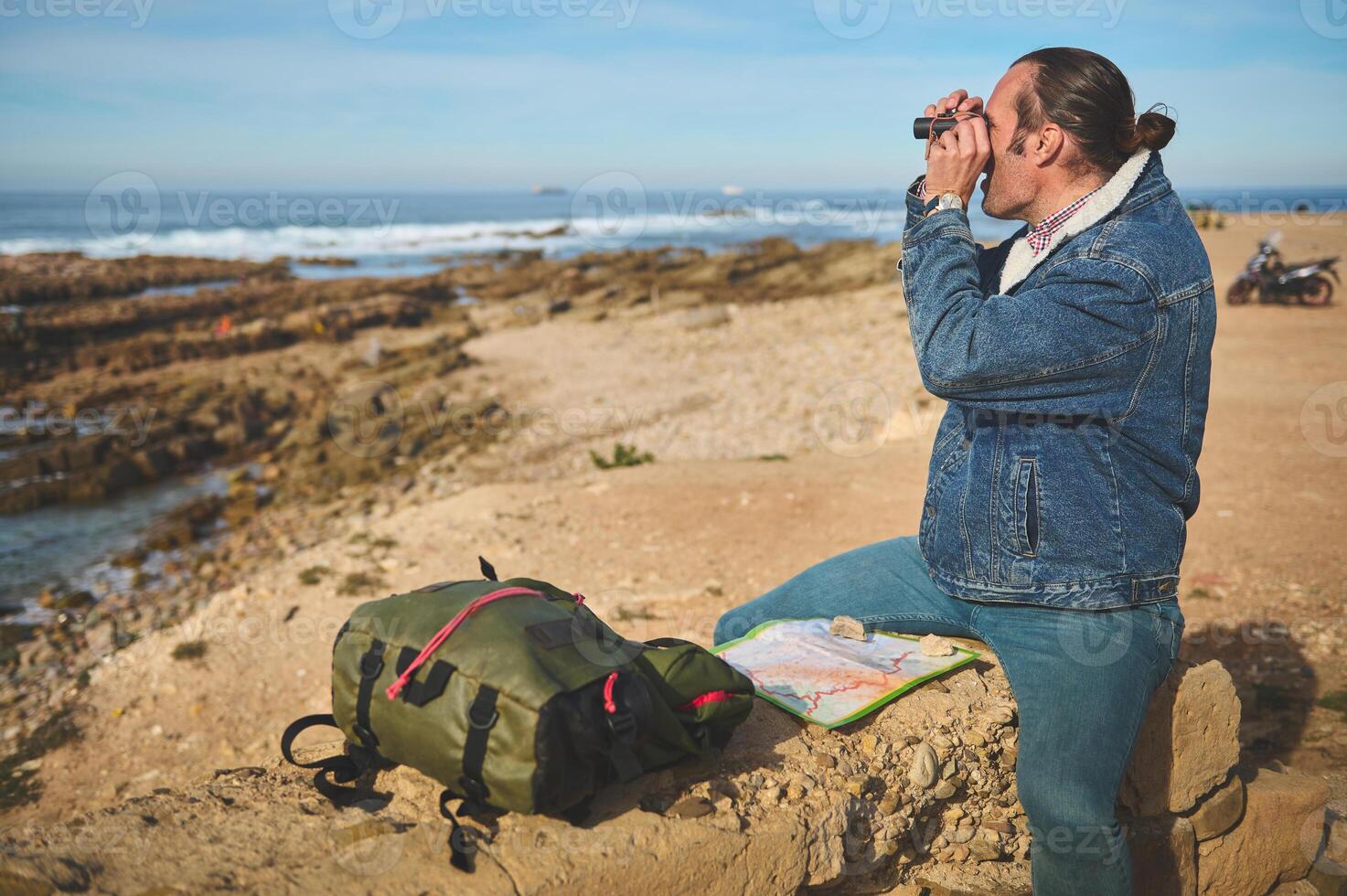 Young male tourist traveler sitting on a rock by sea near his backpack, looking into the distance through his binoculars photo