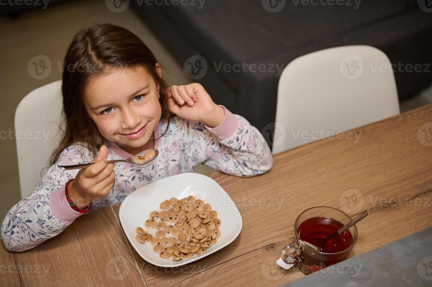 adorable pequeño niño niña lindo sonriente mirando a cámara, sentado a mesa y tomando su delicioso sano desayuno foto