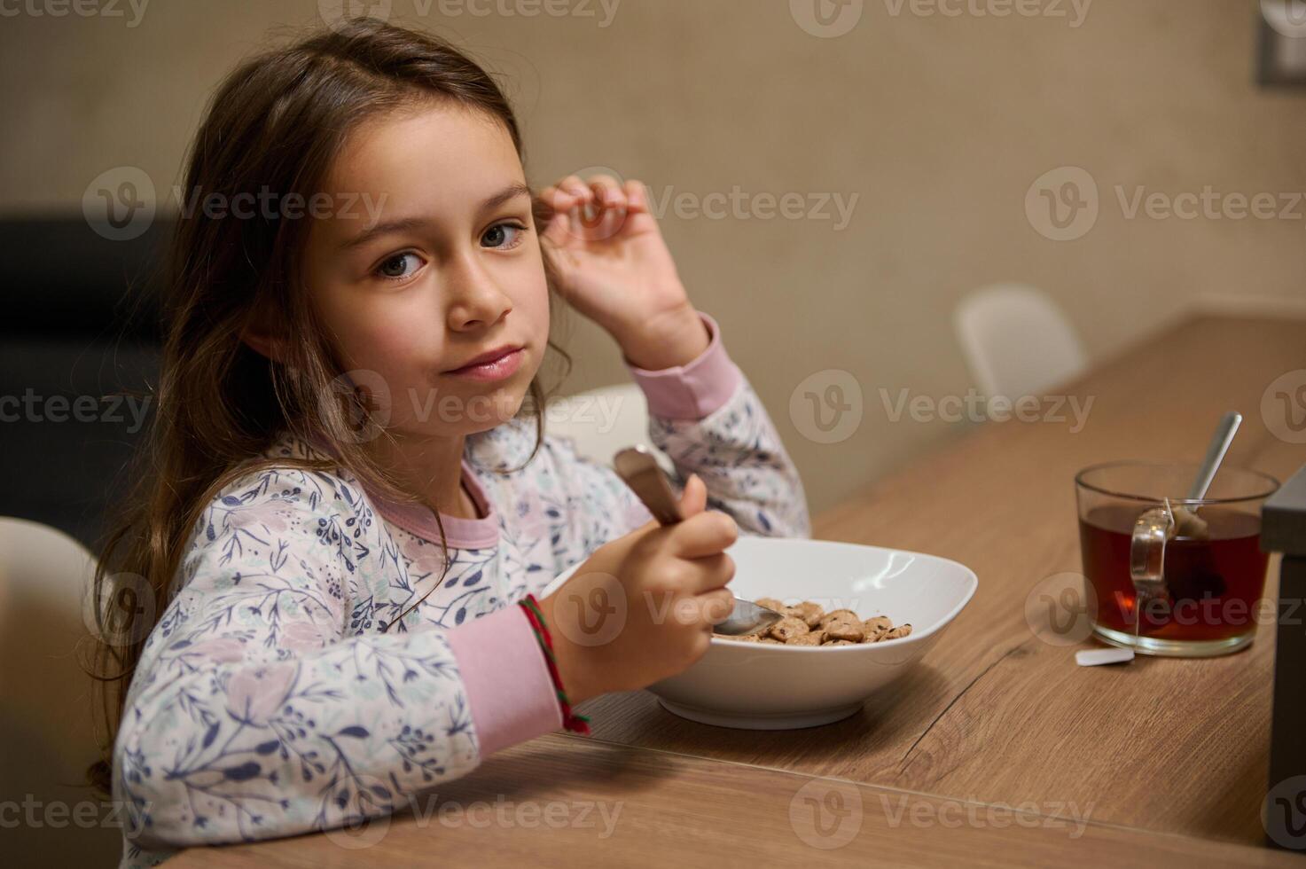 Authentic portrait of a beautiful Caucasian little child girl enjoys eating cereal for morning breakfast with appetite. photo