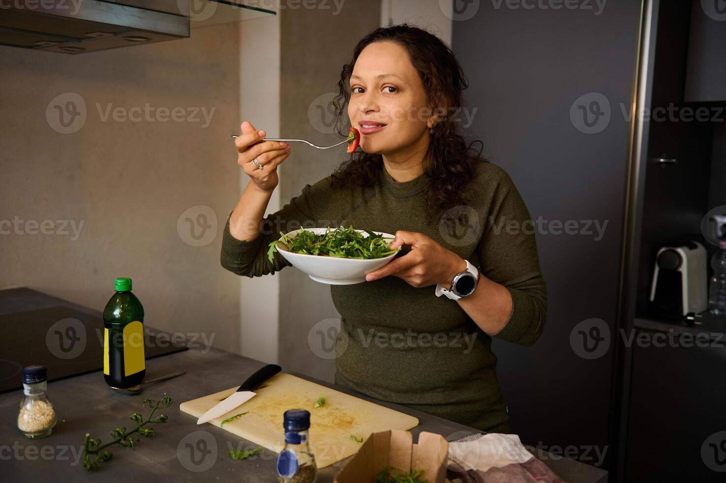 Pretty young woman standing at kitchen table, holding a fork with salad near her face, smiles cutely looking at camera photo