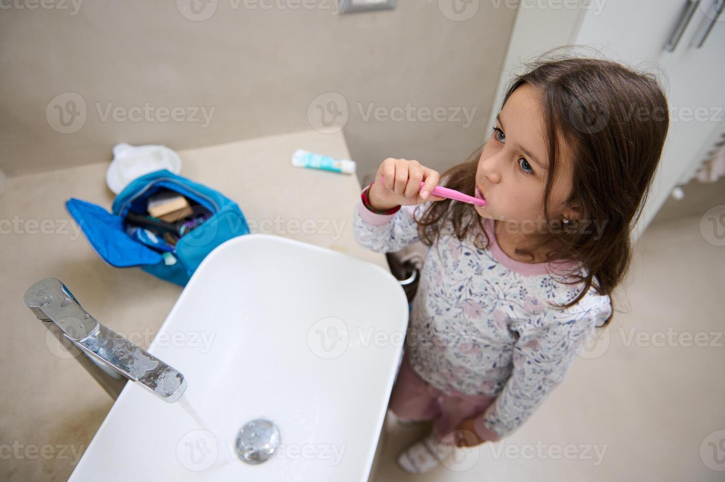 View from above of a child girl brushing teeth, standing at white washbasin in the home bathroom. Dental hygiene concept photo