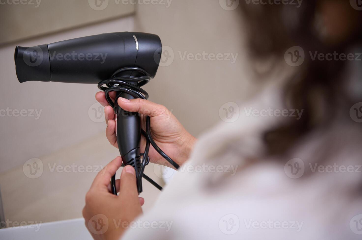 Selective focus on a stylish black hair dryer in the hands of a young brunette woman in white waffle bathrobe. photo