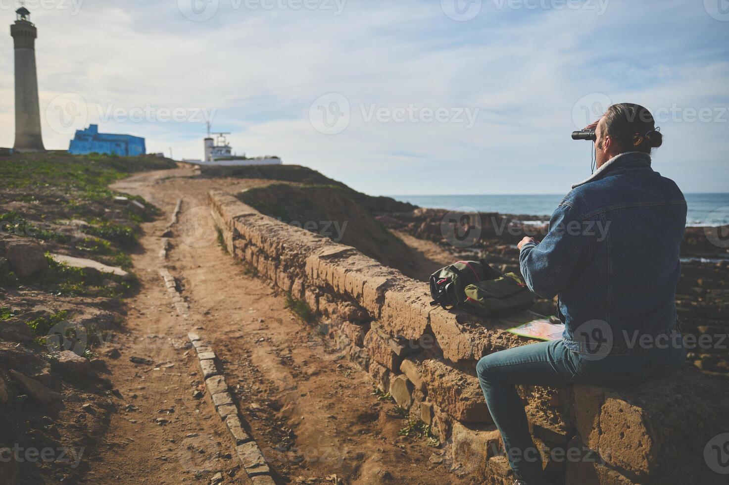 Rear view of a traveler man looking into the distance through binoculars, sitting by sea against lighthouse background photo