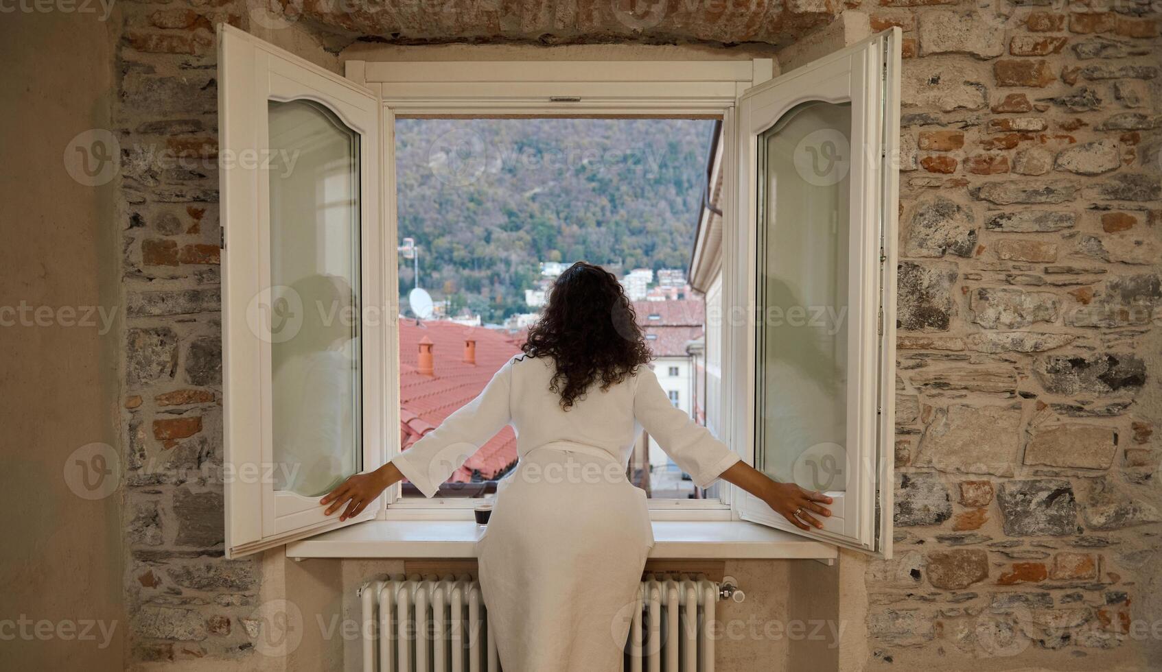 Rear view woman in bathrobe, opens white shutters and windows after waking up in the morning, admiring the city view photo