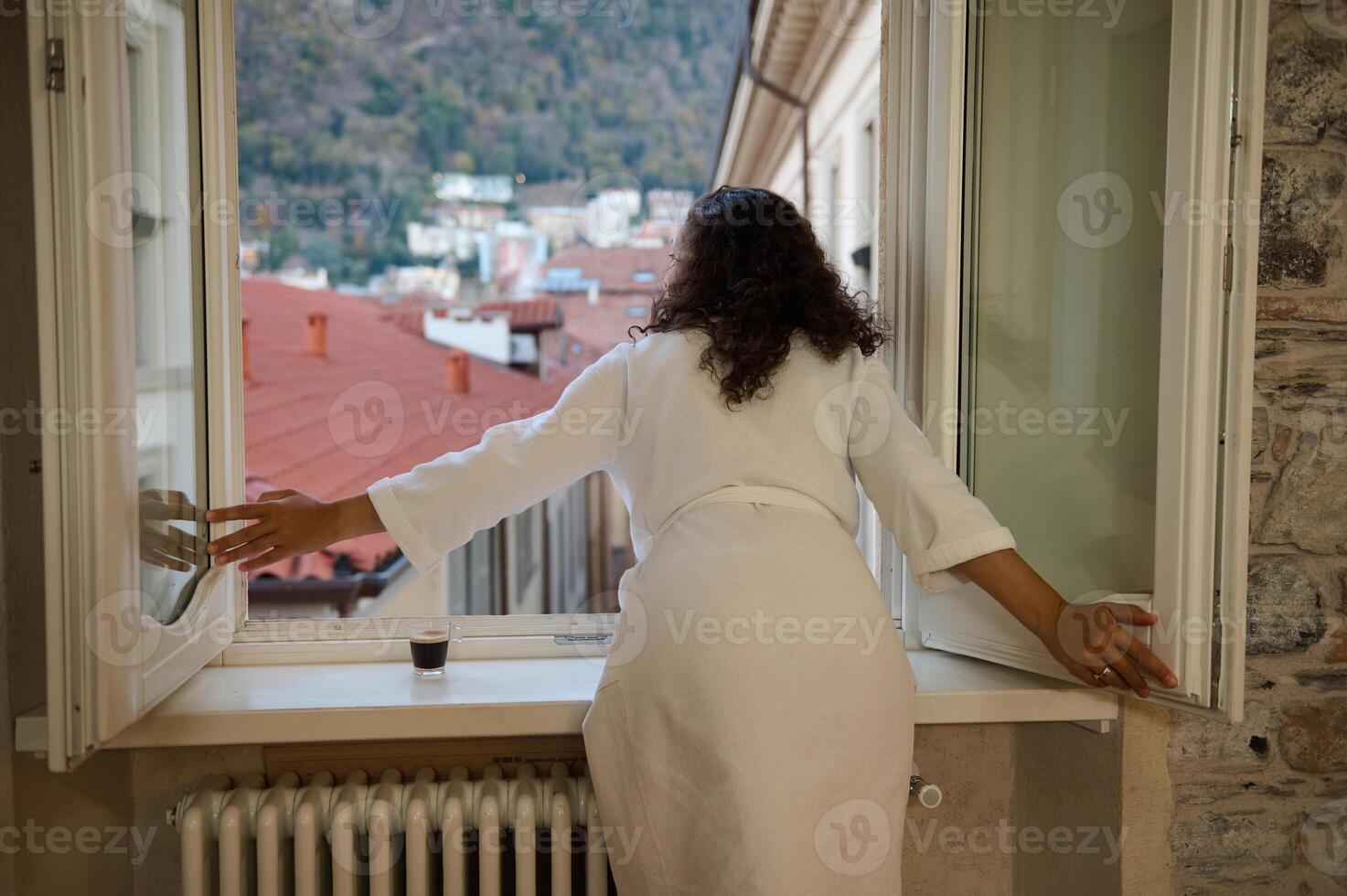 Back View of a pretty woman in white bathrobe, opening windows in the living room, overlooking beautiful Como city photo