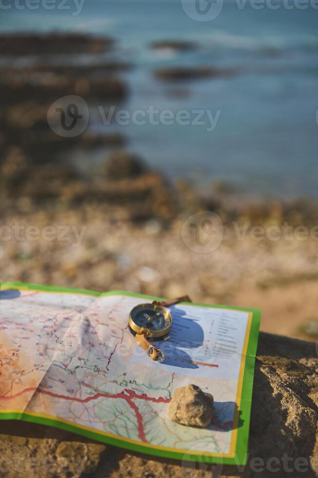 Close-up of an old golden compass and geographic map on a rock over seascape background. Trip. Tourism. Travel concept. photo