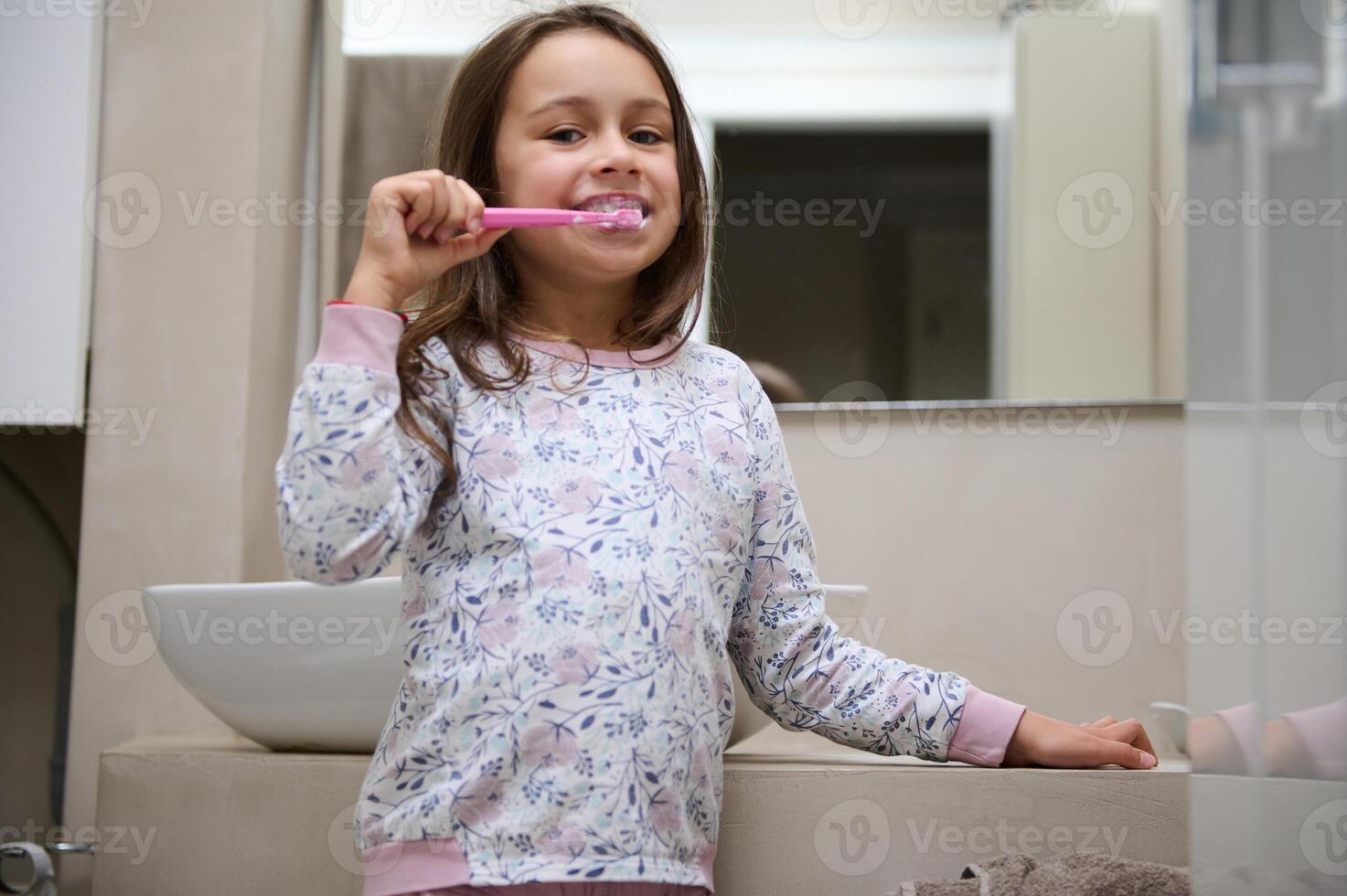 Little kid girl brushing her teeth, standing in the gray minimalist home bathroom, smiling looking at camera. Oral care photo