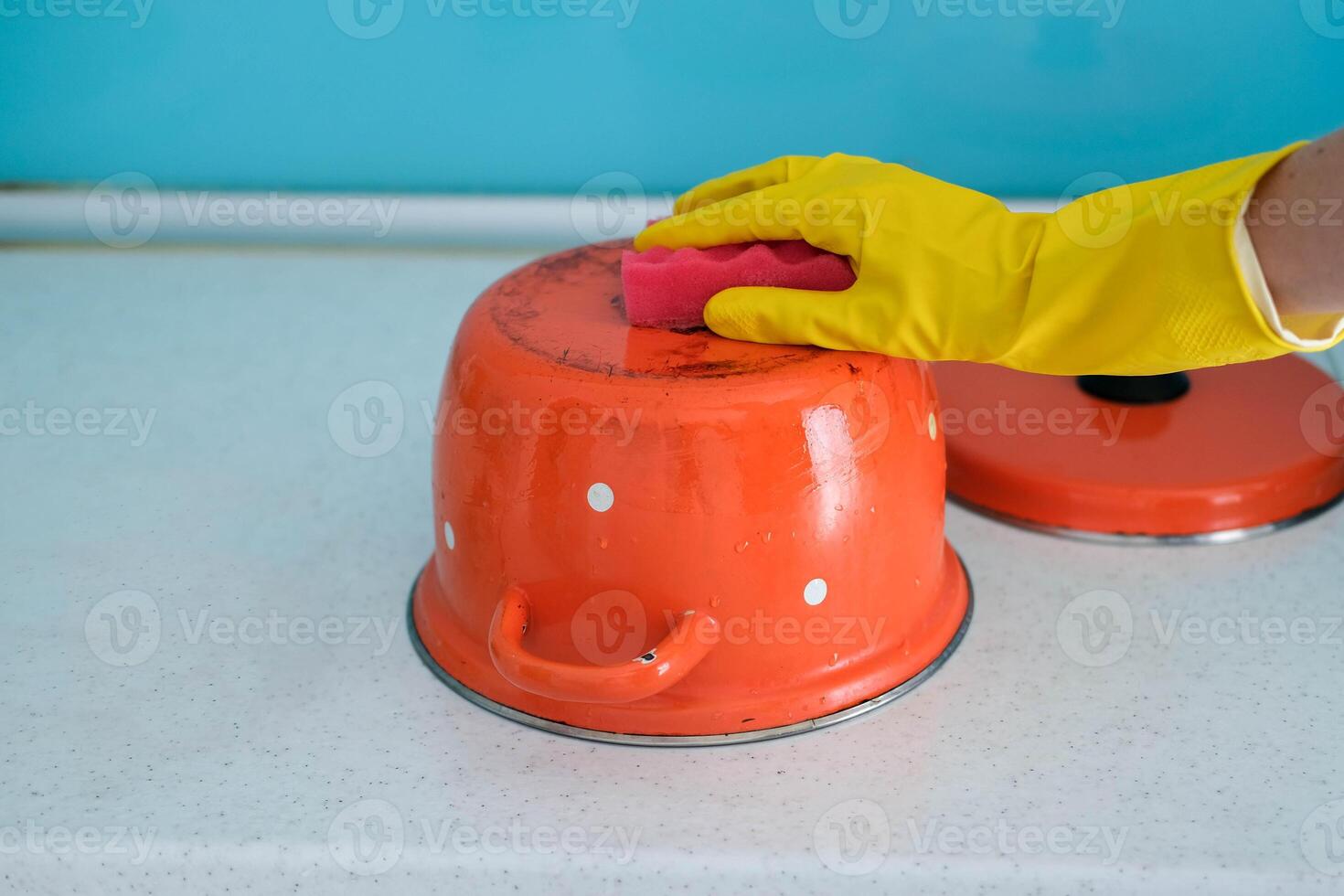 A woman in a hand in a yellow rubber glove washes a dirty red pan with a sponge. photo