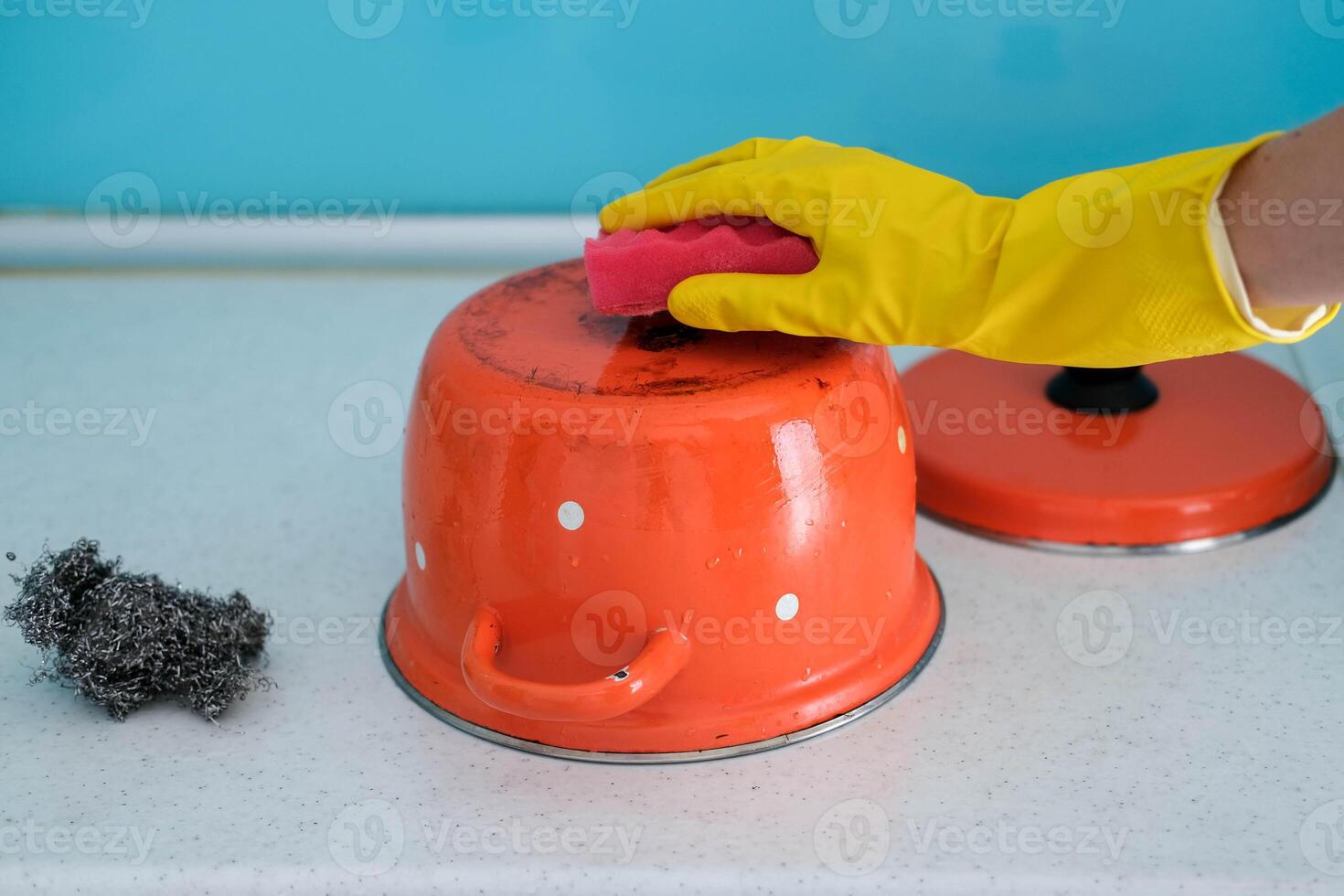 A woman in a hand in a yellow rubber glove washes a dirty red pan with a sponge. photo