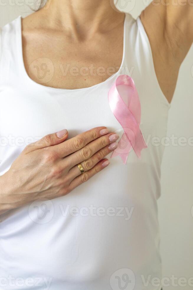 Shot of the woman against the white wall in the white top with pink ribbon, as a symbol of a breast cancer awareness, performing self examination of the breasts, looking for abnormalities. Concept photo