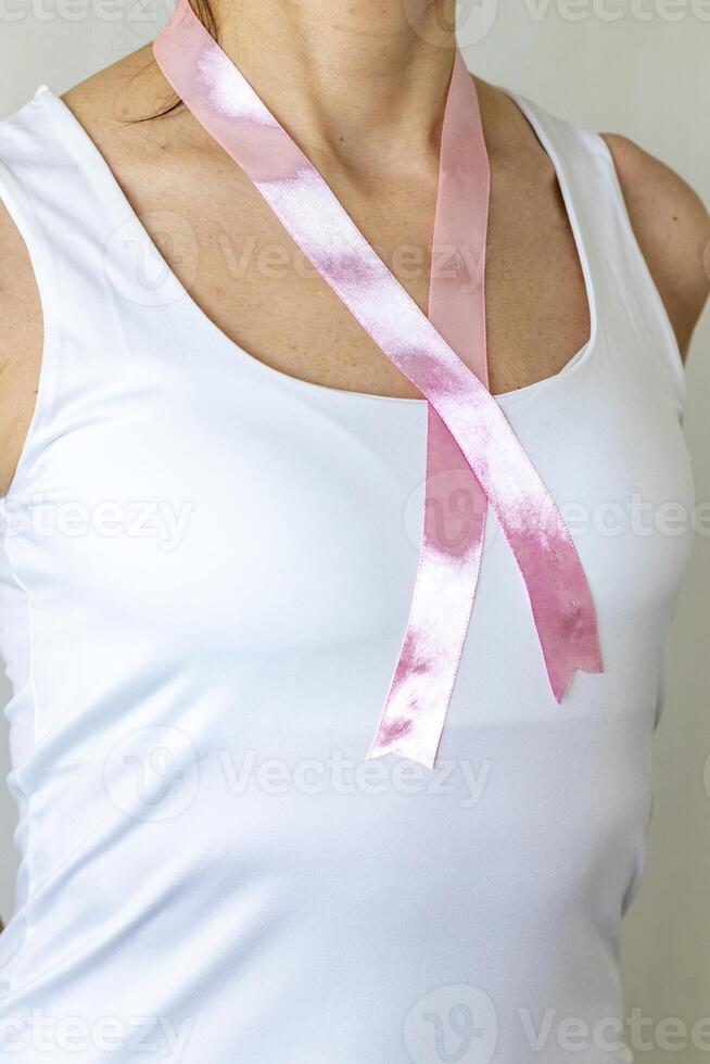 Shot of the woman in the white top against the white wall, with pink ribbon on her neck as a symbol of breast cancer awareness. Concept photo