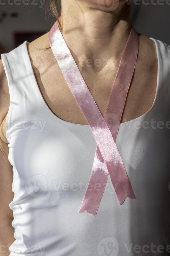 Shot of the woman in the white top against the white wall, with pink ribbon on her neck as a symbol of breast cancer awareness. Concept photo