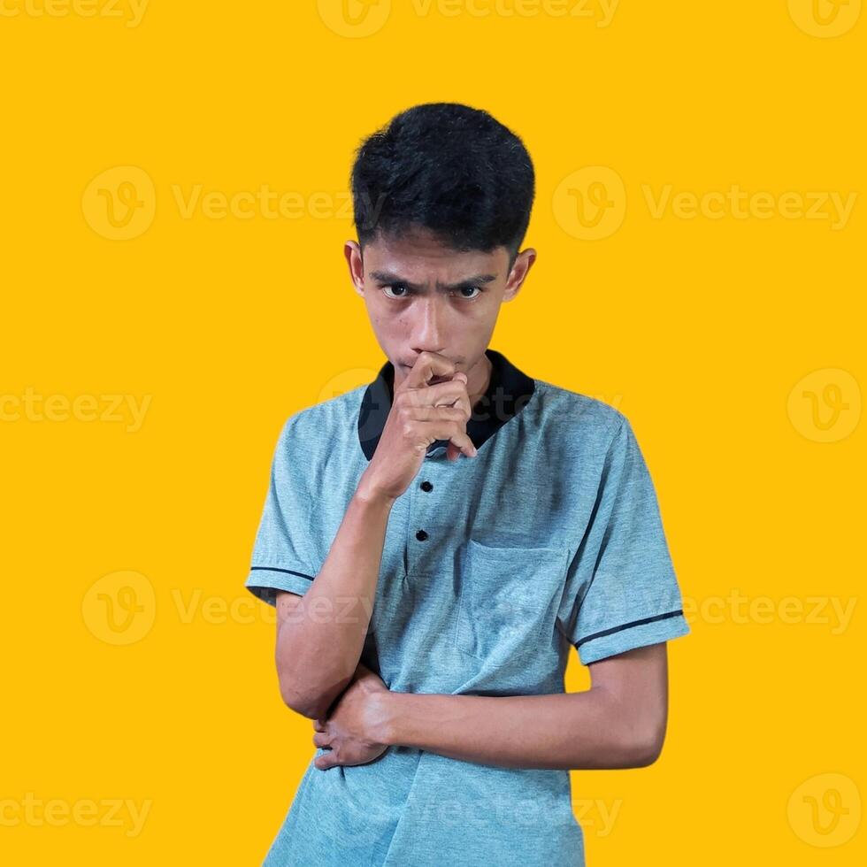 joven pensativo asiático hombre participación mano en barbilla mirando pensativo, vistiendo gris camiseta en amarillo antecedentes. foto