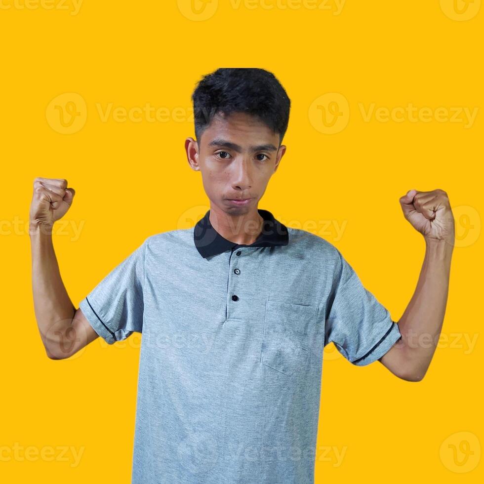 Excited Asian man wearing gray t-shirt showing strong movement raising arms, yellow background. photo