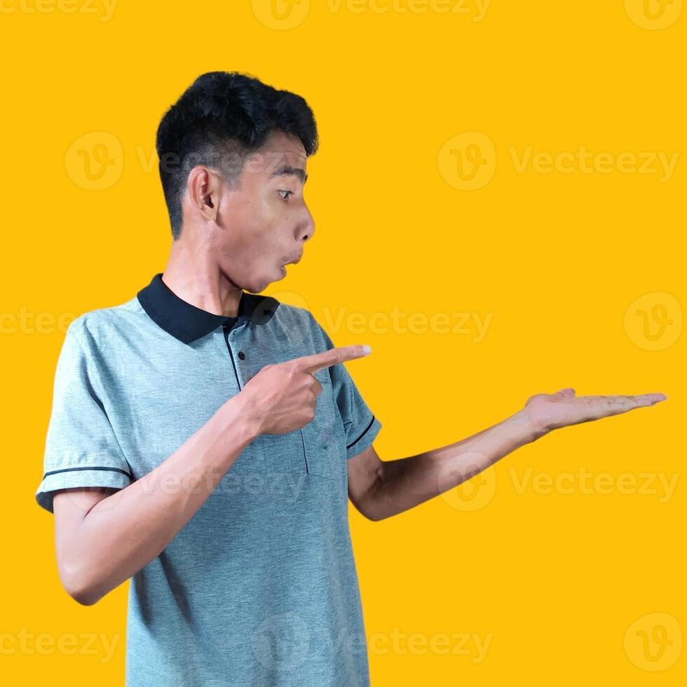 Excited Asian man wearing gray t-shirt pointing at copy space on the side, isolated on yellow background. photo