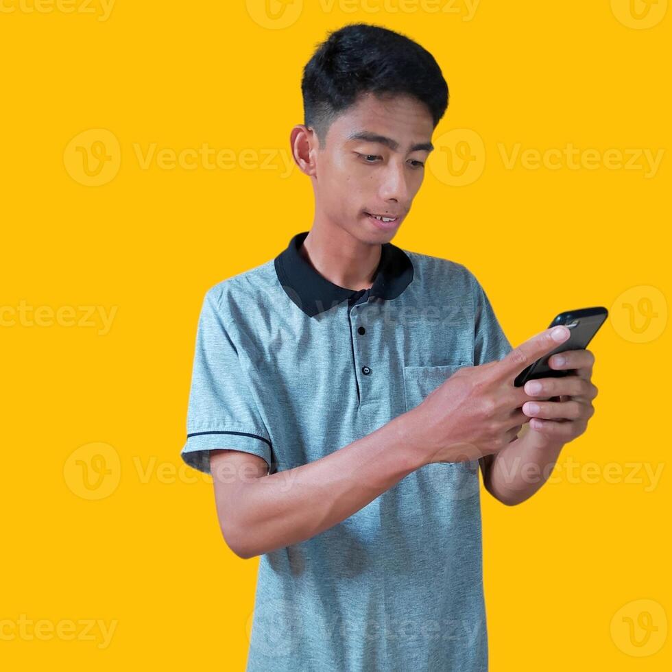 Excited Asian man wearing gray t-shirt pointing to copy space next to holding smart phone, isolated on yellow background. photo