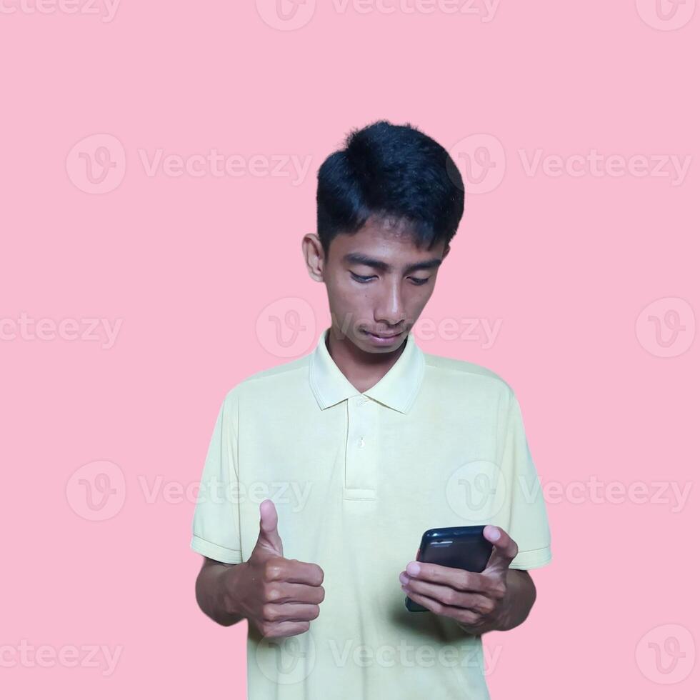 Excited Asian man wearing yellow t-shirt pointing to copy space next to holding smart phone, isolated on pink background. photo