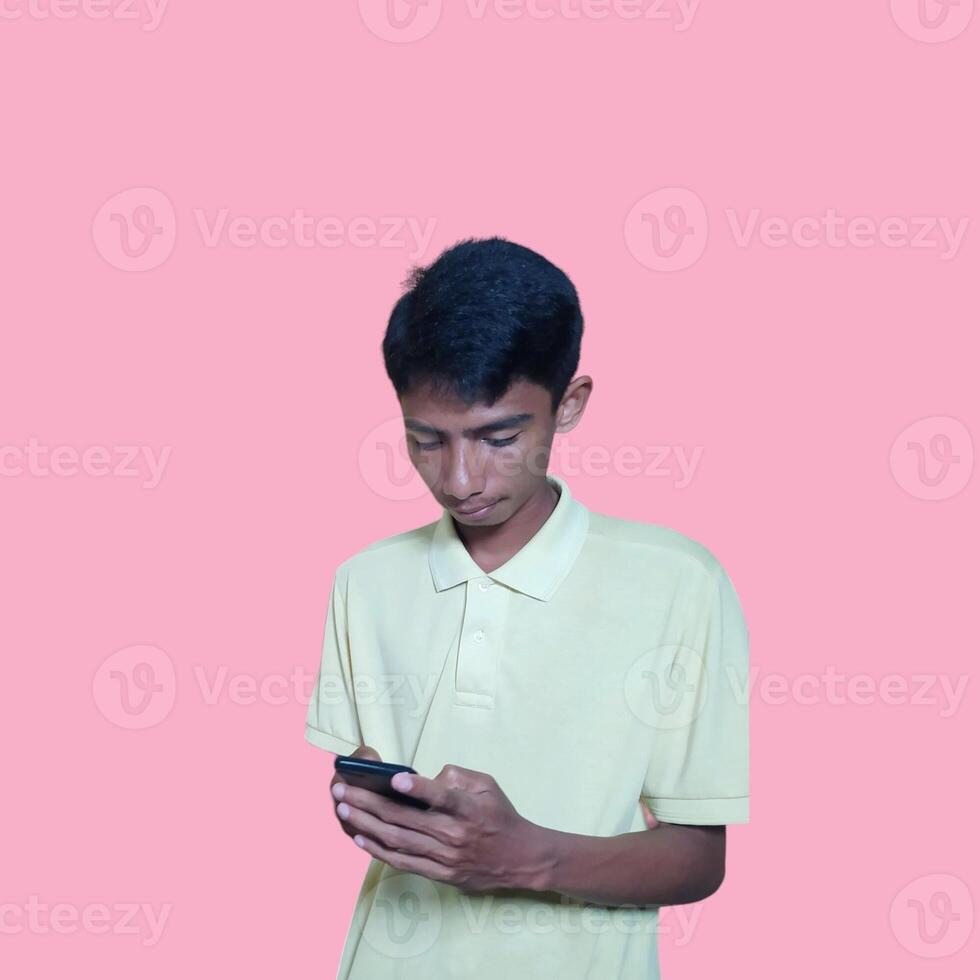 Excited Asian man wearing yellow t-shirt pointing to copy space next to holding smart phone, isolated on pink background. photo