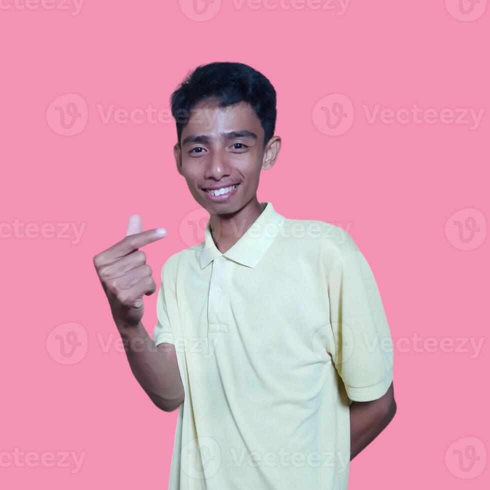 Young Asian man feeling happy and romantic forming heart gesture, wearing yellow t-shirt on pink background. photo