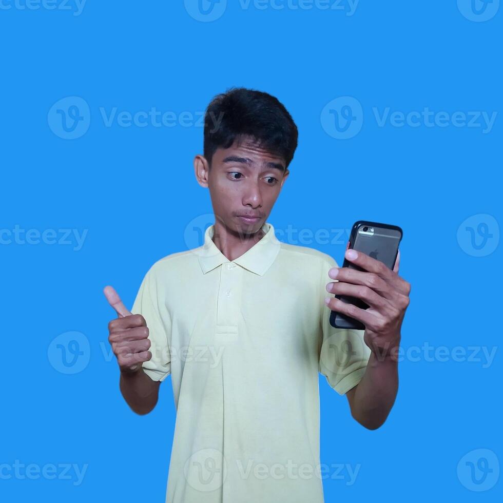 Young Asian man surprised looking at smart phone wearing yellow t-shirt, blue background. photo