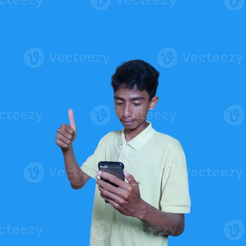 joven asiático hombre sorprendido mirando a inteligente teléfono vistiendo amarillo camiseta, azul antecedentes. foto