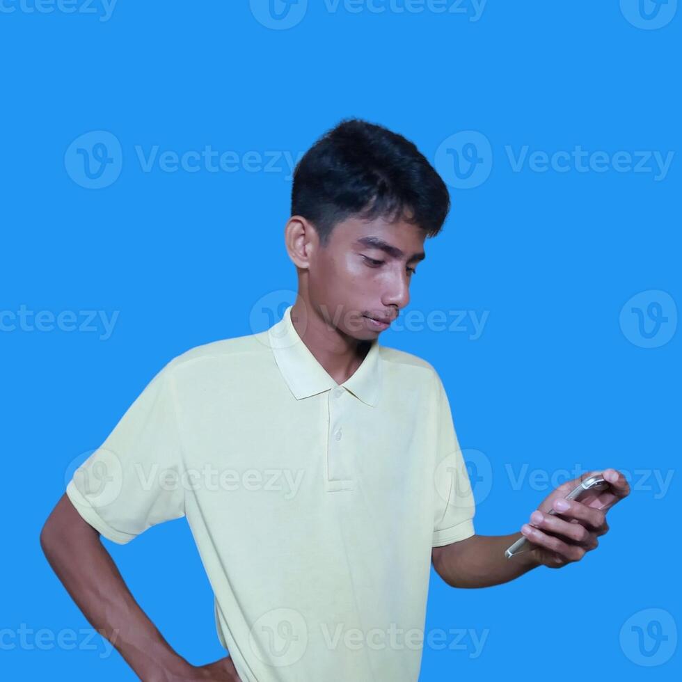 joven asiático hombre sorprendido mirando a inteligente teléfono aislado azul antecedentes. vistiendo un amarillo camiseta foto