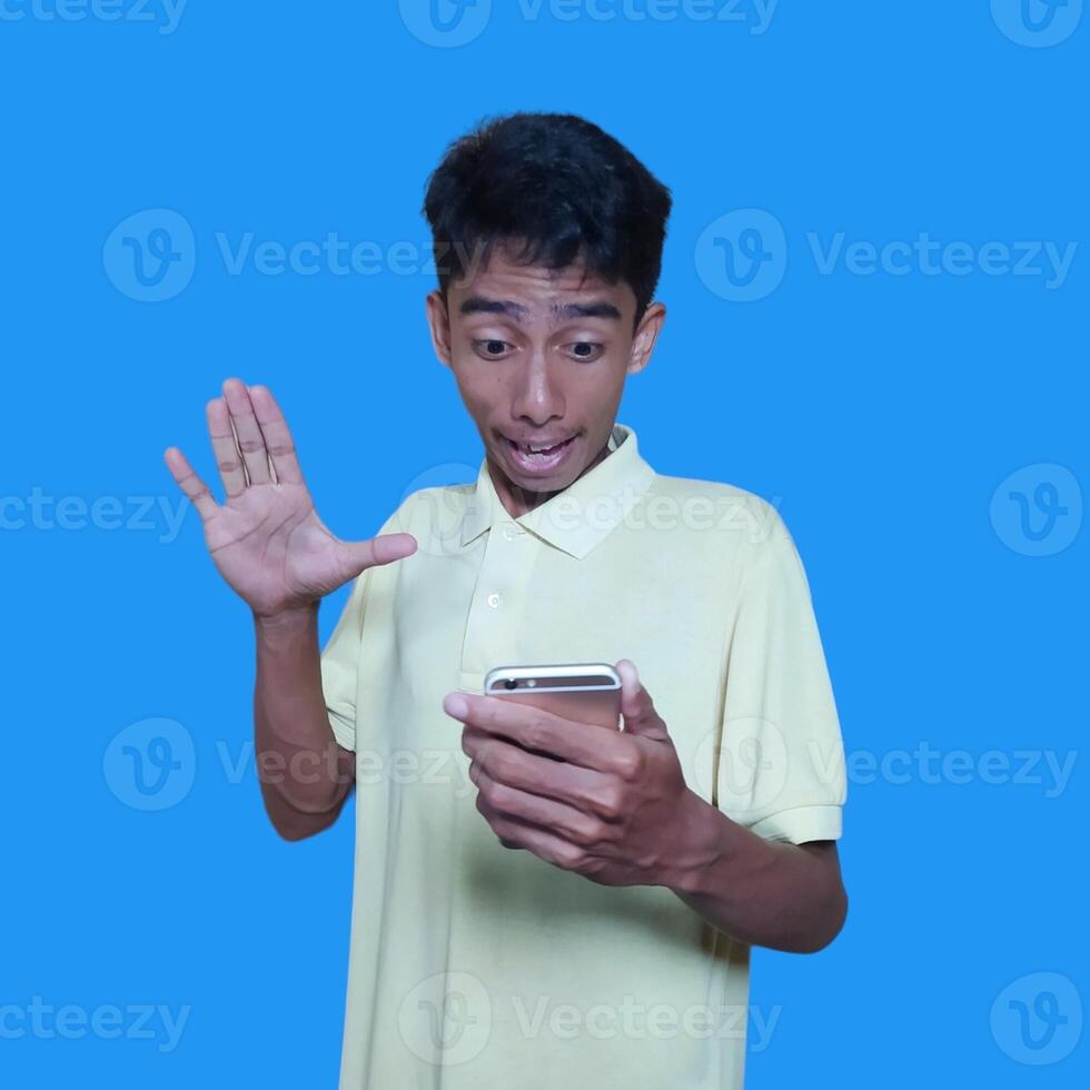 joven asiático hombre sorprendido mirando a inteligente teléfono aislado azul antecedentes. vistiendo un amarillo camiseta foto