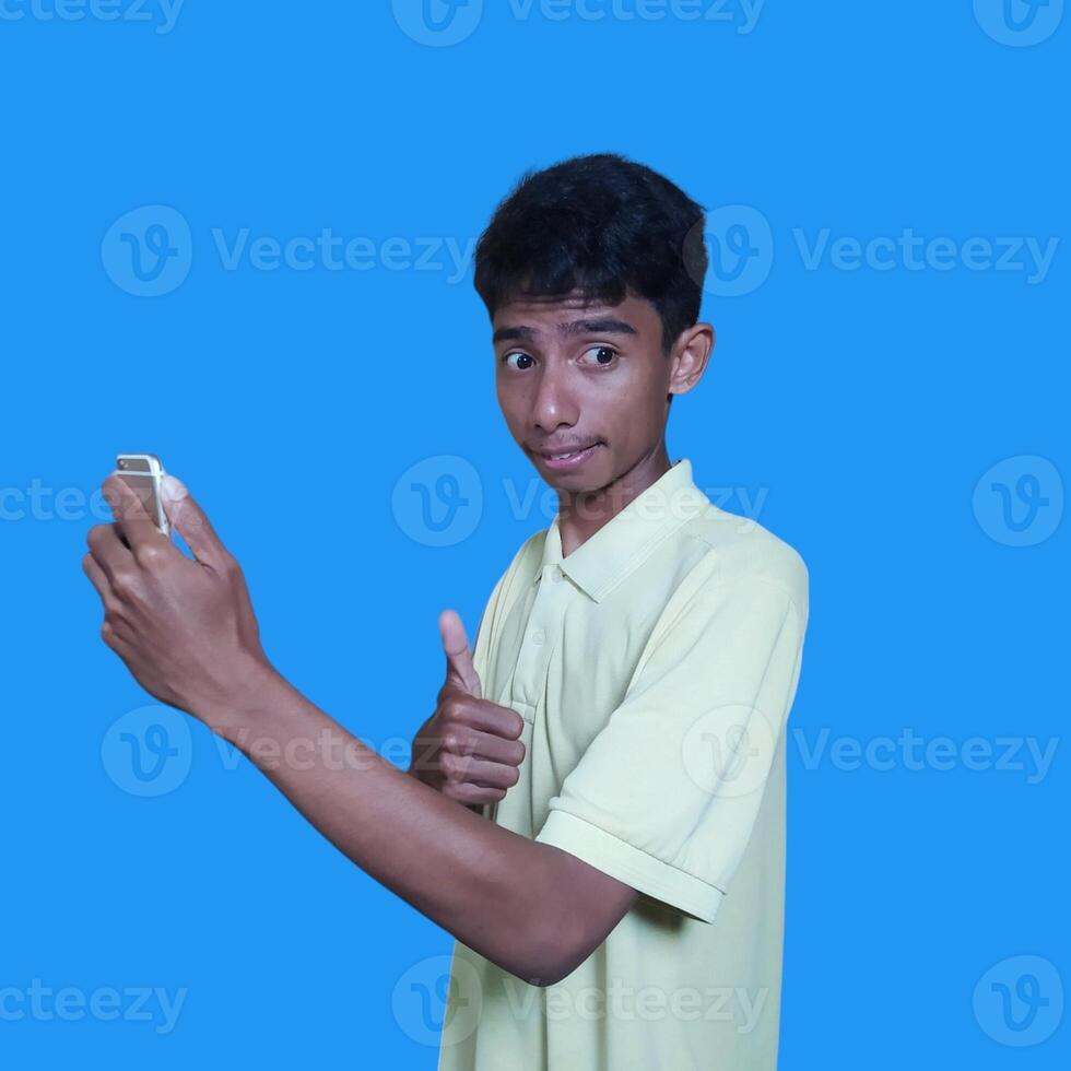 joven asiático hombre sorprendido mirando a inteligente teléfono aislado azul antecedentes. vistiendo un amarillo camiseta foto
