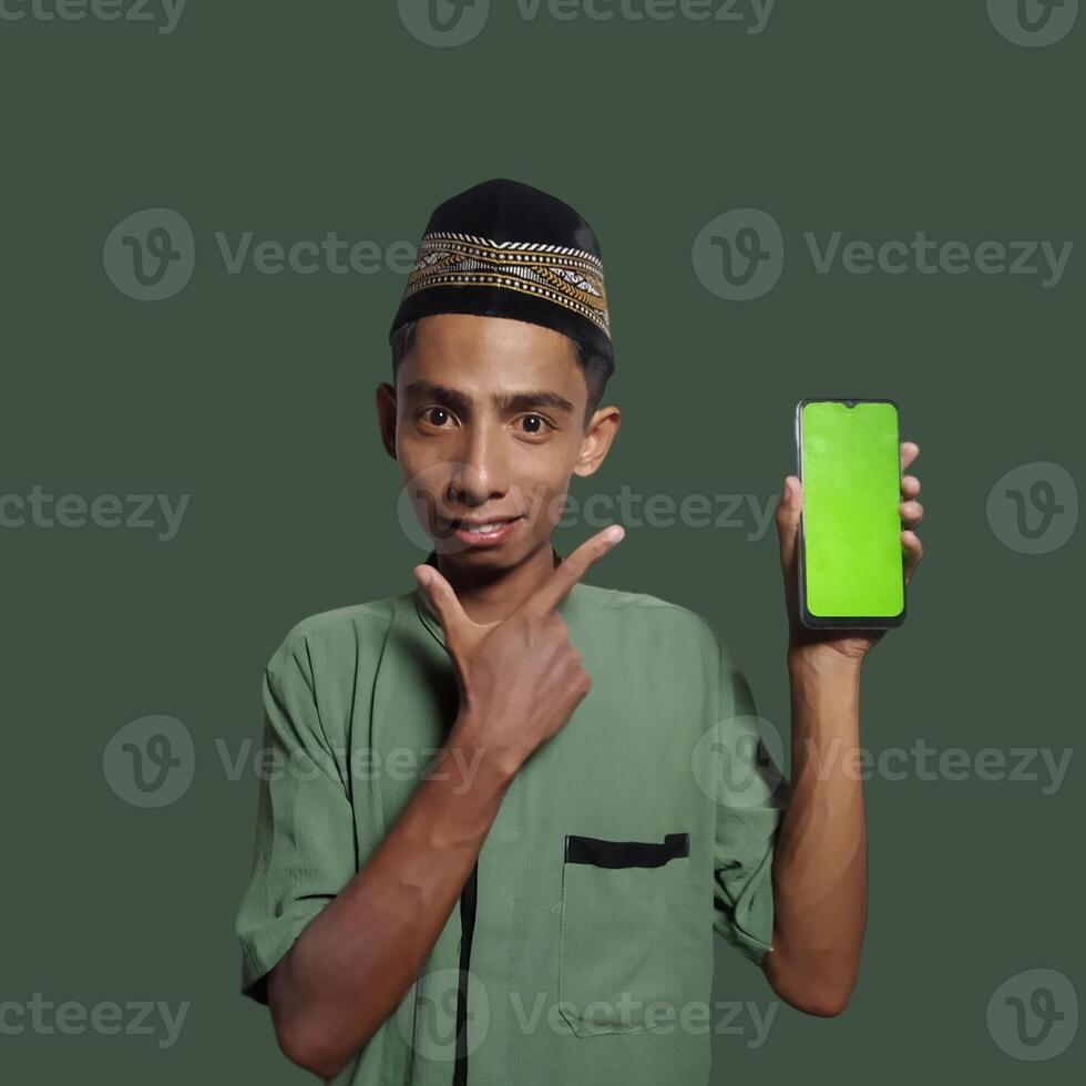 A Young Asian Muslim Man wearing a Skullcap while showing a mobile phone screen isolated on a green background. photo