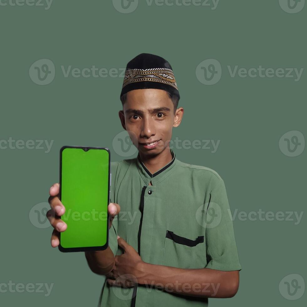 A Young Asian Muslim Man wearing a Skullcap while showing a mobile phone screen isolated on a green background. photo