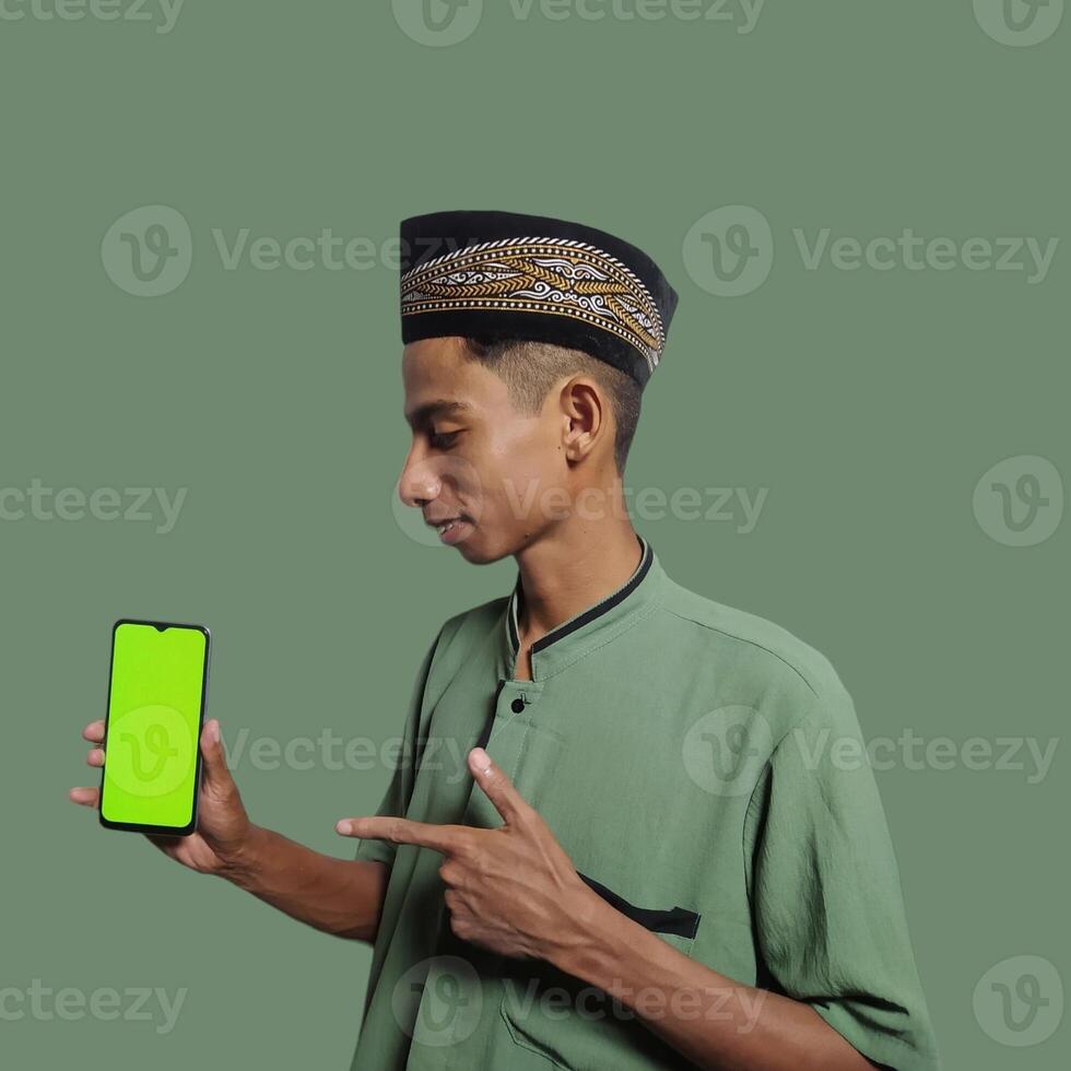 A Young Asian Muslim Man wearing a Skullcap while showing a mobile phone screen isolated on a green background. photo