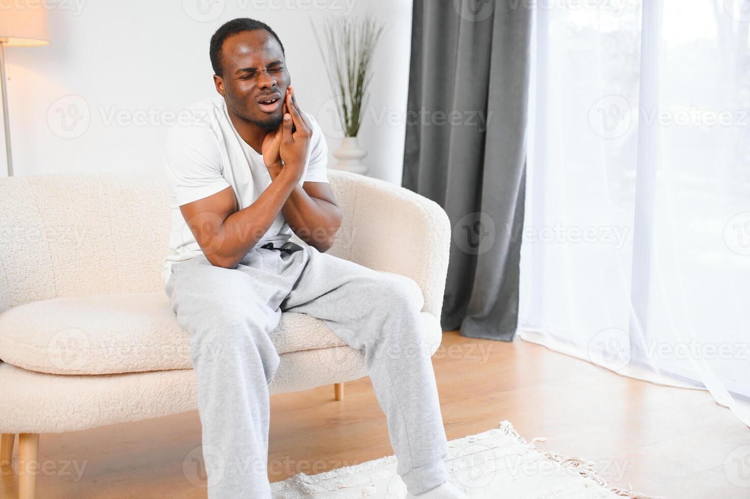 Close-up photo of a young African-American man suffering from a severe toothache at home on the couch. He holds his hand to his cheek, grimacing in pain