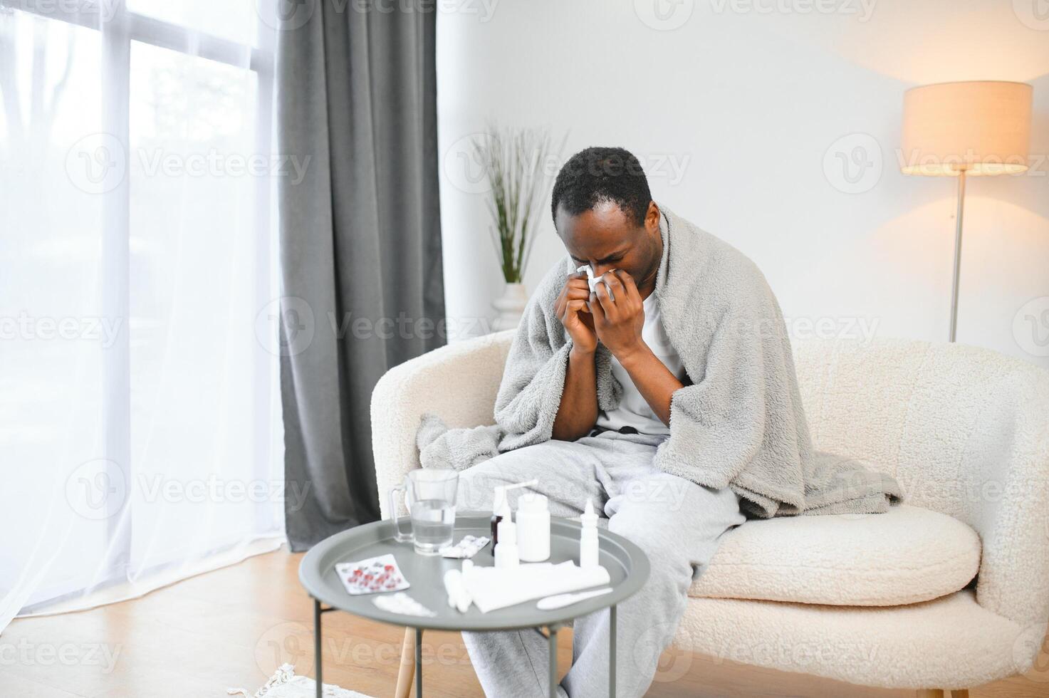 Portrait of a sick young african american man wrapped in a blanket on the sofa. Guy with seasonal flu or cold feel unhealthy with influenza and runny nose at home photo