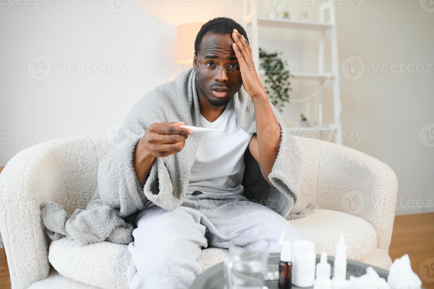 Fever. Portrait of sick african american man with thermometer suffering from flu symptoms sitting wrapped in blanket in bed at home photo