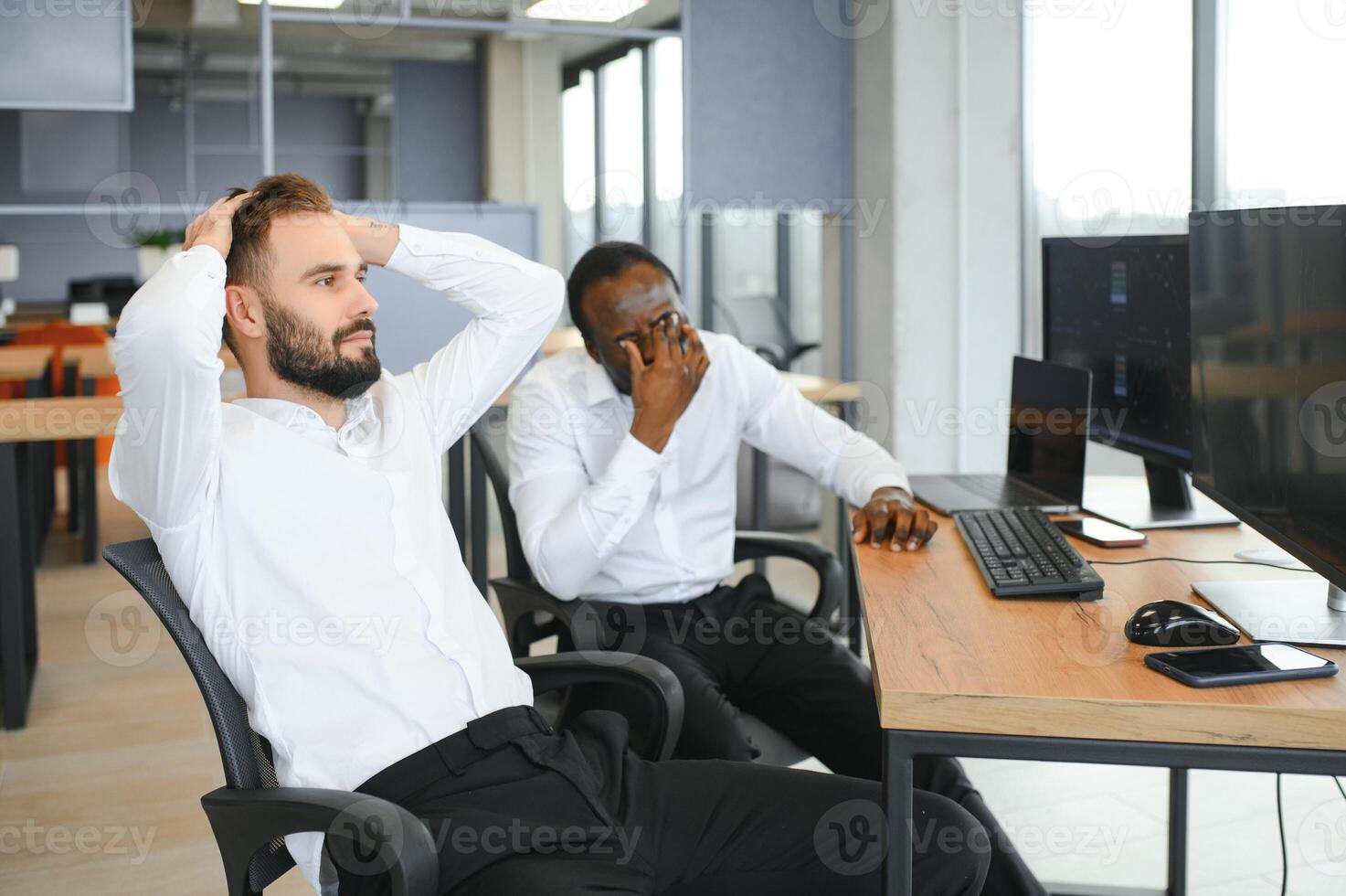 Two diverse colleagues traders talking to each other, sitting in the office in front of multiple computer screens. Stock trading, people, business concept photo