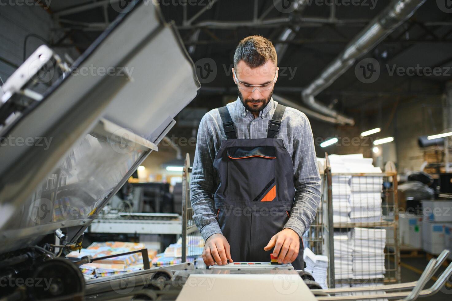 Printing house interior with modern offset print machine and operator in working uniform checking quality and controlling process of print photo
