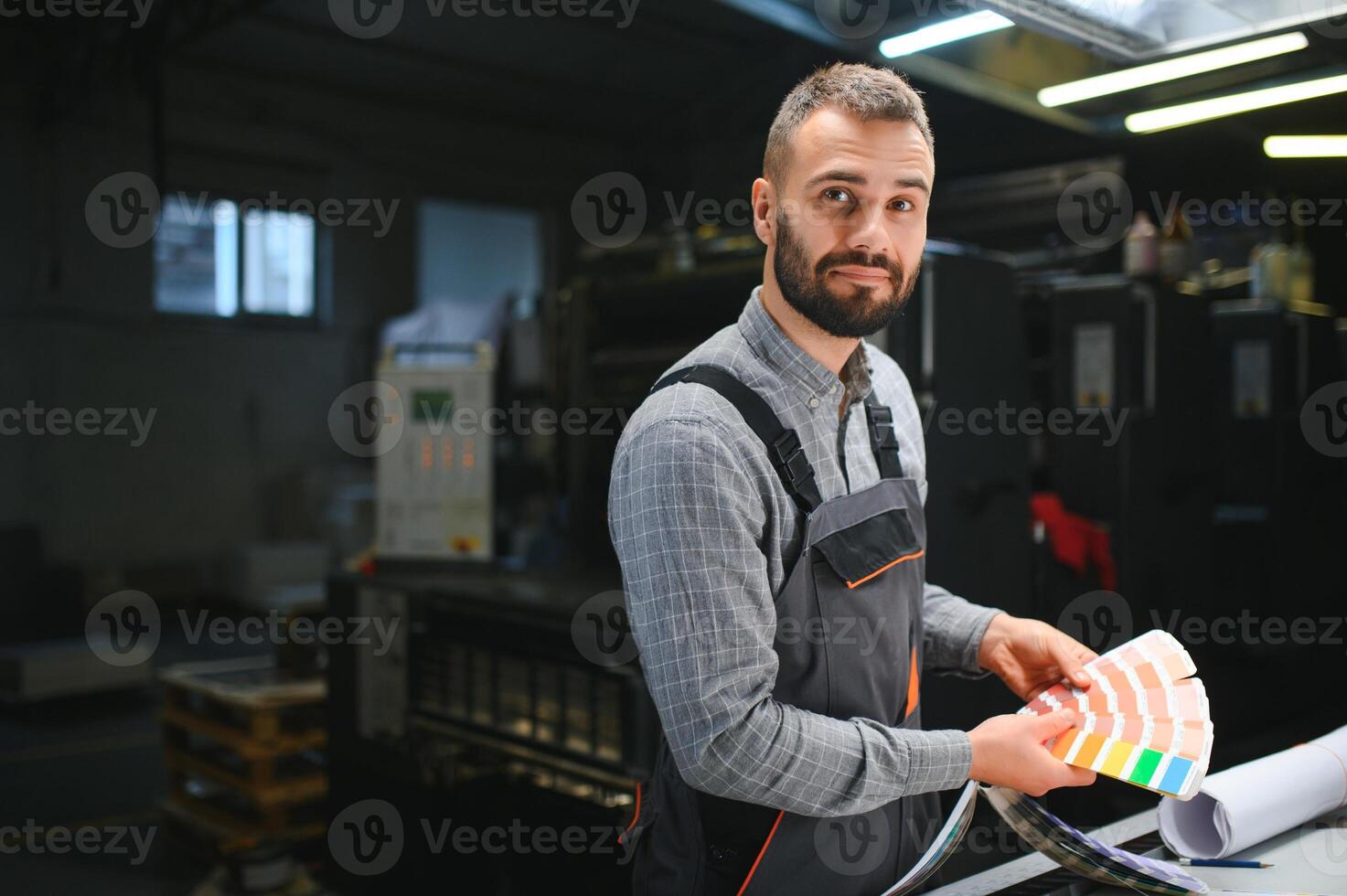 Printing house interior with modern offset print machine and operator in working uniform checking quality and controlling process of print photo