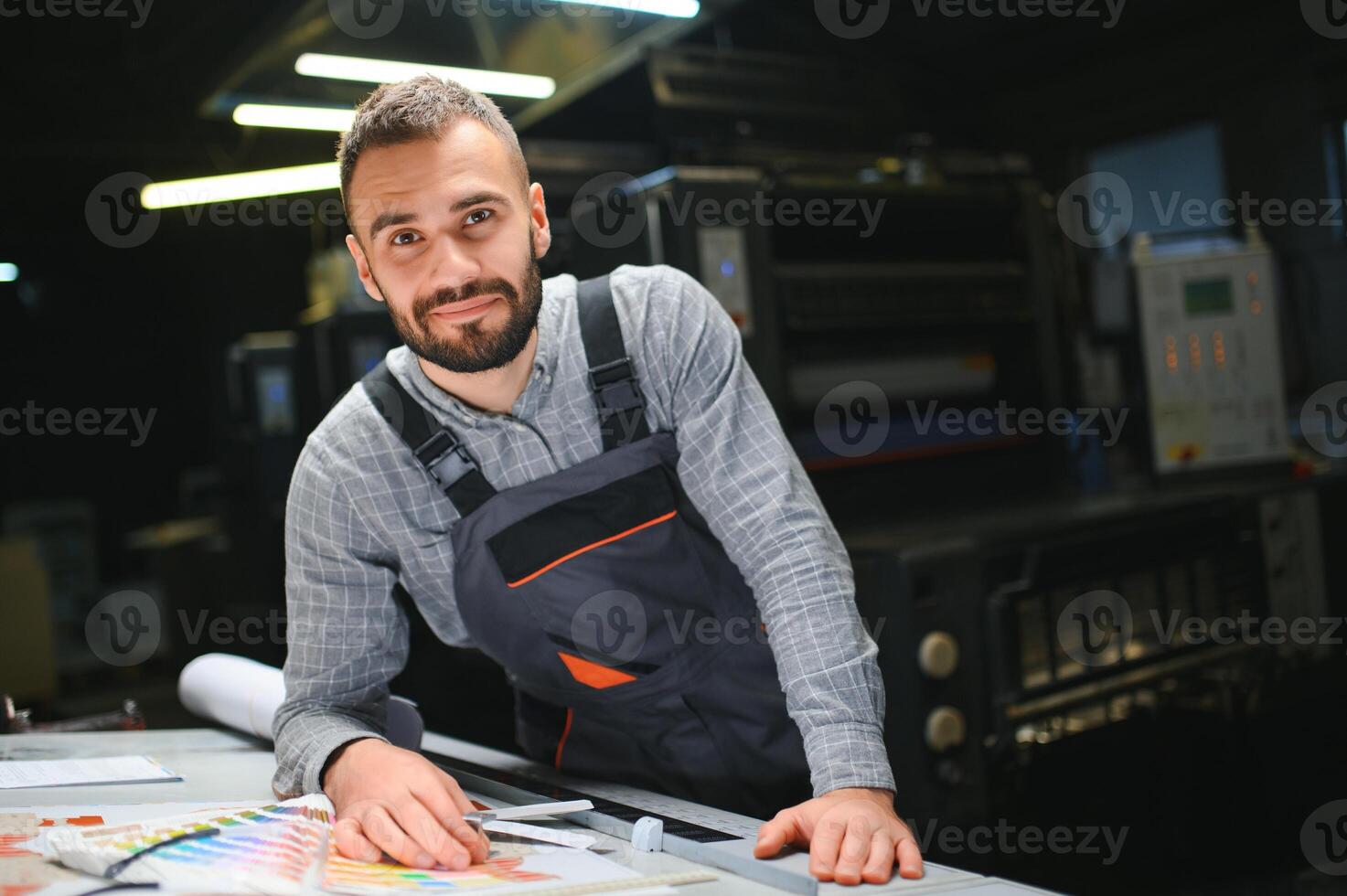 Printing house interior with modern offset print machine and operator in working uniform checking quality and controlling process of print photo